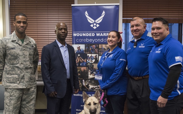A group of five people and a service dog pose together in front of a 'Wounded Warrior' Air Force banner at what appears to be a recruitment or awareness event. The group includes a uniformed Air Force military member, a civilian in a suit, and three individuals wearing blue Air Force-branded polo shirts. A German Shepherd service dog wearing a pink harness sits in the center of the group.