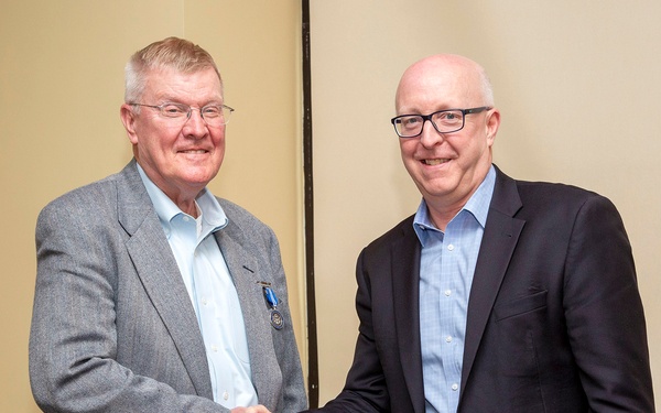 Two men in business casual attire are shaking hands and smiling in what appears to be an indoor setting, likely an office or meeting room. The man on the left is wearing a gray blazer and has a blue medal or pin affixed to his lapel, suggesting a formal recognition or award ceremony. The overall tone of the image is celebratory and professional.
