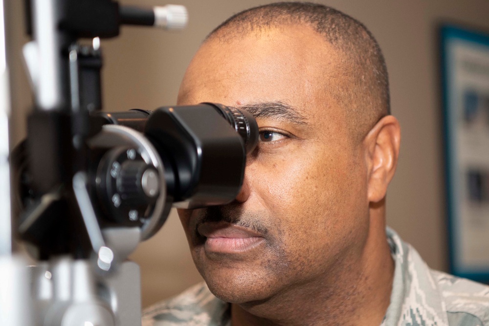 A man in military camouflage uniform is undergoing an eye examination using a slit lamp or similar ophthalmic diagnostic device. The image is a close-up shot focusing on his face and the medical equipment positioned in front of his eye. The setting appears to be a medical or optometry clinic.