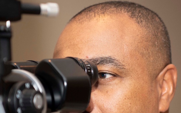 A man in military camouflage uniform is undergoing an eye examination using a slit lamp or similar ophthalmic diagnostic device. The image is a close-up shot focusing on his face and the medical equipment positioned in front of his eye. The setting appears to be a medical or optometry clinic.