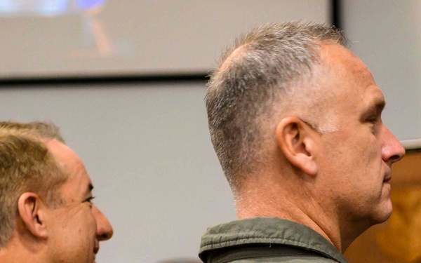 Two military personnel are seated in what appears to be a briefing room or auditorium, facing toward a speaker at a podium in the background. The individual on the left wears a MultiCam/OCP camouflage uniform, while the individual on the right wears an olive drab flight suit with a patch visible on the shoulder. A projection screen is visible in the background, suggesting an official military presentation or ceremony is underway.