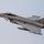A military jet fighter is captured in flight against an overcast gray sky. The aircraft displays Italian Air Force markings, including a roundel visible on the fuselage, identifying it as an Eurofighter Typhoon. The aircraft appears to be in a slight banking maneuver with landing gear partially visible.