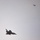 Two military jet aircraft are visible in flight against an overcast, grey sky. The foreground aircraft is clearly identifiable as a delta-wing fighter jet with a camouflage paint scheme, while a second aircraft is visible at a greater distance in the upper right. The image appears to have been taken from the ground looking upward.