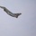 A military fighter jet is captured in flight against an overcast gray sky. The aircraft appears to be a Eurofighter Typhoon, identifiable by its delta wing configuration and canard foreplanes. The jet is banking at an angle, suggesting a climbing or turning maneuver.
