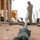 A U.S. Marine in desert MARPAT camouflage uniform is handling a flagpole or mast assembly on sandy ground at what appears to be a military training or forward operating base. The low-angle perspective emphasizes the length of the metal pole, with its base mechanism visible in the foreground. The surrounding environment includes wooden structures, concrete barriers, coiled rope, and various equipment scattered on the ground.