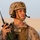 A U.S. Marine in desert combat gear is holding a military radio (handheld transceiver) while wearing a combat helmet with communications headset. The soldier appears to be on duty in a desert environment, actively monitoring or preparing to communicate via radio. A name tape reading 'MUNAFO' and a blood type tag 'O POS' are visible on the body armor.