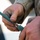 A close-up photograph of a person's hands wearing MARPAT (Marine pattern) digital camouflage uniform, manipulating what appears to be a cleaning rod or similar tubular tool. The hands are visibly dirty/grimy, suggesting active field work or maintenance activity. The image is tightly cropped to focus on the hands and the tool being assembled or disassembled.