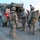 A group of approximately five soldiers in full combat gear are gathered around the rear ramp of a military armored vehicle, likely a Stryker, marked with a red cross indicating a medical variant. The scene appears to take place at dusk or dawn in an arid, desert-like training or operational environment. The soldiers are equipped with helmets, body armor, backpacks, and weapons.