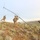 Three military personnel in camouflage uniforms are working together to erect a tall antenna mast on a hillside in an arid, scrubland terrain. The soldiers appear to be setting up a communications antenna system, with guy-wires visible extending from the top of the mast. The landscape is characterized by sparse sagebrush and rolling hills under an overcast sky.