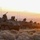 A column of military wheeled armored vehicles, identifiable as Stryker Infantry Carrier Vehicles (ICVs), are positioned in a line on arid, scrubland terrain at either sunrise or sunset. The vehicles are equipped with weapon stations, multiple radio antennas, and external gear/equipment. Unit markings are partially visible on at least one vehicle, including what appears to read 'A-33'.