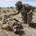 Two U.S. Army soldiers in full combat uniforms and gas masks are engaged in what appears to be a CBRN (Chemical, Biological, Radiological, Nuclear) training exercise in an open field. One soldier is kneeling and attending to another soldier who is lying on the ground, simulating a casualty scenario. Military tents and vehicles are visible in the background, indicating a field training environment.