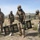 Four military personnel wearing gas masks and combat gear are conducting what appears to be a CBRN (Chemical, Biological, Radiological, Nuclear) training exercise in an arid field environment. One soldier in the center is carrying a folding mesh stretcher or litter, while others stand nearby in various uniforms including desert camouflage and OCP/MultiCam patterns. Military tents and equipment are visible in the background.