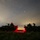 A nighttime military scene showing two armored wheeled vehicles, likely Stryker APCs, positioned in an open field or scrubland under a star-filled sky. A prominent red light source illuminates the ground between the two vehicles, suggesting active operations or a signaling device. Personnel are visible atop both vehicles, appearing to be on watch or conducting operations.