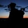 A silhouetted military figure is photographed against a twilight sky with orange and blue gradient hues at dusk or dawn. The soldier is equipped with night vision goggles mounted on a helmet and carries a rifle over their shoulder. The dark foreground terrain and distant hills are barely visible against the colorful horizon.