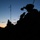 A silhouetted military soldier is positioned on elevated terrain during twilight or dawn conditions. The soldier appears to be in a tactical observation or overwatch position, equipped with helmet-mounted devices and a radio antenna visible against the sky. A second silhouetted figure with optical equipment is visible in the middle distance.
