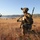 The image shows a group of military personnel conducting a field operation across an open grassland terrain with a body of water and rolling hills visible in the background. The foreground soldier is in full combat gear including helmet-mounted night vision devices, body armor, and is carrying an assault rifle while in a kneeling security position. Multiple soldiers are dispersed across the field in what appears to be a tactical patrol or training exercise.