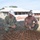 Two U.S. Navy personnel in camouflage uniforms and hard hats are crouching down to inspect a rebar/reinforcement steel grid at an active construction site. The individual on the left is wearing a white hard hat with a rank insignia and a name tag reading 'CAREY', while the individual on the right wears a tan hard hat and sunglasses. A building under construction or renovation is visible in the background, surrounded by orange safety fencing.