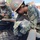 Military personnel in camouflage uniforms are engaged in construction work at an active job site. The foreground worker wears a hard hat with a safety sticker and yellow safety glasses while working with rebar and wooden formwork. Multiple service members are visible in the background performing various construction tasks.