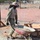 A military service member in camouflage uniform and tan hard hat is operating a red wheelbarrow filled with gravel or aggregate material at an active construction site. The site is cordoned off with orange safety fencing, with wooden formwork visible nearby. The background shows a dry, open area with motorcycles parked and civilians present.