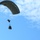 A military-style ram-air parachute in olive/gray coloring is descending through a partly cloudy blue sky. Suspended beneath the canopy is a person along with what appears to be a cargo bundle or equipment package. This appears to be a military airborne operation or supply drop exercise.