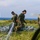A group of military personnel in camouflage uniforms are gathered on a grassy coastal field, working to gather and pack a deployed parachute. The scene appears to be a post-jump recovery operation, with the ocean visible in the background. At least four service members are actively involved in handling the parachute lines and canopy.