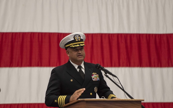 A U.S. Navy officer in dress blues is delivering a speech at a podium bearing the USS America (LHA 6) seal. A second naval officer is seated in the background, smiling. The event appears to be a formal military ceremony, likely a change of command, conducted in front of a large American flag backdrop.