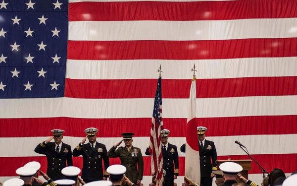 A formal military ceremony is taking place inside a large indoor venue, likely a hangar, with a massive American flag as the backdrop. Personnel from multiple branches — including U.S. Navy and U.S. Marine Corps — are rendering salutes, with a Japanese flag also visible alongside the U.S. flag, suggesting a joint or bilateral military event. The scene is consistent with a change of command, commissioning, or official ceremony.