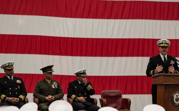 A formal U.S. military ceremony is taking place in front of a large American flag backdrop. A Navy officer is speaking at a podium bearing what appears to be a ship's crest, while four seated officers — including Navy personnel and one Marine — observe from behind. An audience of enlisted sailors wearing white dixie cup caps fills the foreground.