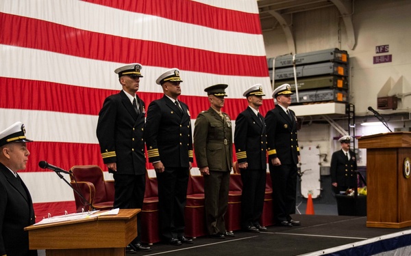 A formal U.S. military ceremony, likely a change of command or commissioning event, taking place inside what appears to be a naval vessel's hangar bay. A naval officer stands at a podium addressing the gathering, while five officers — including Navy personnel and one Marine in service uniform — stand at attention on a raised platform. A large American flag serves as a backdrop, and the stage is draped in red, white, and blue bunting.