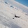 An aerial refueling operation is taking place above a dense cloud layer. A large tanker aircraft is connected via a refueling boom or drogue to a smaller fighter jet below it. The image is taken from above or alongside the aircraft, with blue sky visible in the upper right corner.