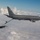 A U.S. Air Force KC-46 Pegasus tanker aircraft (tail number 76026) is conducting an aerial refueling operation with an F-16 Fighting Falcon fighter jet above a cloud layer. The boom refueling system is visibly extended and connected between the two aircraft. Both aircraft are painted in gray military livery and flying in clear blue sky conditions.