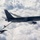 The image shows a U.S. Air Force aerial refueling operation in flight above a cloud layer. A large tanker aircraft, identifiable as a KC-46 Pegasus, is connected via a refueling boom to a smaller F-16 Fighting Falcon fighter jet. Both aircraft are painted in dark gray livery and are flying in close formation during the fuel transfer.