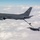 A U.S. Air Force KC-46 Pegasus tanker aircraft (tail number 76026) is conducting an aerial refueling operation with an F-16 Fighting Falcon fighter jet. The boom refueling system is visibly extended and connected between the two aircraft. Both aircraft are flying above a dense cloud layer under overcast skies.