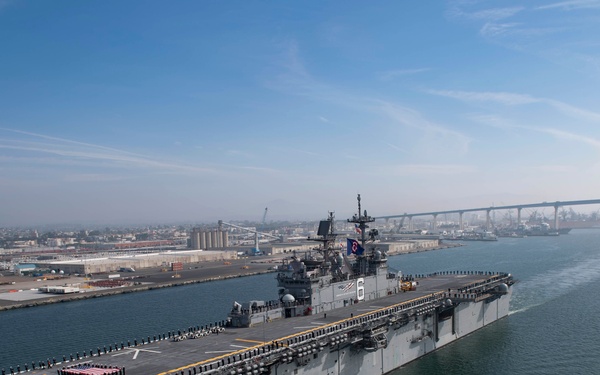 An aerial view of a large U.S. Navy amphibious assault ship underway in what appears to be San Diego Bay, with the Coronado Bridge visible in the background. The ship's flight deck is lined with sailors in dress uniform, suggesting a formal departure or arrival ceremony. Industrial port facilities and the San Diego skyline are visible along the shoreline.