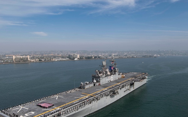 An aerial photograph of a large US Navy amphibious assault ship underway in what appears to be San Diego Bay, with the city skyline visible in the background. The ship's flight deck is lined with sailors in dress uniform, suggesting a formal departure or arrival ceremony. The hull number '6' is visible on the island superstructure, consistent with USS America (LHA-6) or a similar Wasp/America-class vessel.