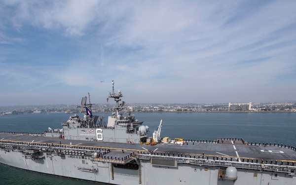 An aerial photograph of a large U.S. Navy amphibious assault ship underway in what appears to be a harbor or bay, with a coastal city visible in the background. The hull number '6' is prominently displayed on the island superstructure, and 'LHA 6' is visible on the hull, identifying this as the USS America. Personnel are lined up along the flight deck edges, suggesting a formal departure or arrival ceremony.
