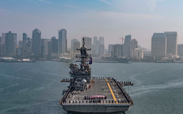 A large U.S. Navy amphibious assault ship is navigating a harbor with a major city skyline visible in the background, consistent with San Diego Bay. The flight deck is lined with sailors in dress uniform, suggesting a formal departure or arrival ceremony. The vessel appears to be an America-class or Wasp-class amphibious assault ship based on its flat-top configuration and hull design.