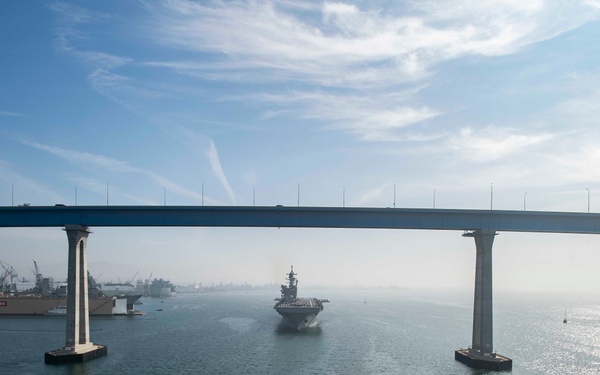 An aerial or elevated view of a large naval aircraft carrier passing beneath a major bridge over a bay or harbor channel. In the background, a shipyard facility with cranes and dry docks is visible, with signage reading 'BAE SYSTEMS'. The scene appears to be a naval vessel either departing or arriving at a port.
