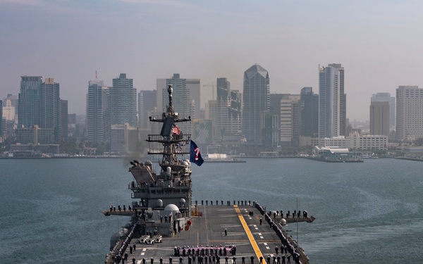 A large U.S. Navy amphibious assault ship is navigating through a harbor with a major city skyline visible in the background, consistent with San Diego, California. The ship's flight deck is lined with sailors in uniform standing at parade rest, suggesting a formal departure or arrival ceremony. Multiple flags, including the American flag, are flying from the ship's mast.