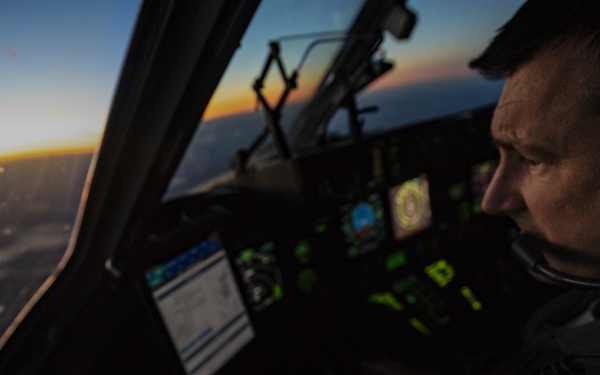 A military pilot is seated in the cockpit of a large aircraft during twilight or dawn conditions, with a vivid orange and blue horizon visible through the windscreen. The pilot is wearing a flight suit with a U.S. flag patch, aviation headset, and is surrounded by illuminated green avionics displays. The scene suggests an active flight mission at high altitude.