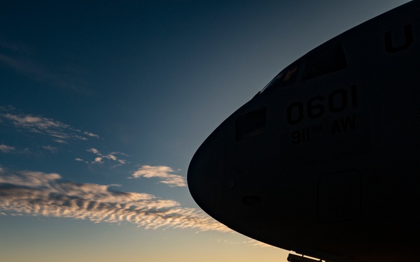 A dramatic silhouette photograph taken at sunset or sunrise on a military airfield, showing a large U.S. military aircraft with the tail number '0601' visible on its fuselage. A single person, likely ground crew or maintenance personnel, stands in silhouette beside the aircraft on the tarmac. The warm golden horizon contrasts sharply with the deep blue sky above, creating a striking compositional effect.