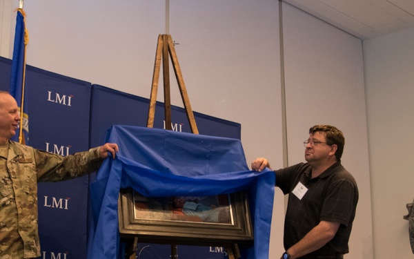 Three individuals are participating in a formal unveiling ceremony at an LMI (Logistics Management Institute) event. Two military personnel in uniform and one civilian are removing a blue cloth to reveal a framed artwork or painting on a wooden easel. The backdrop features repeated LMI branding logos on blue panels.