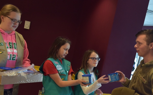 Three Girl Scouts wearing green and tan vests are handing cookies and boxes to a young man in military camouflage attire. The interaction appears to be a cookie donation or sales event taking place inside a building with colorful walls. A second person in civilian clothing is visible in the background.