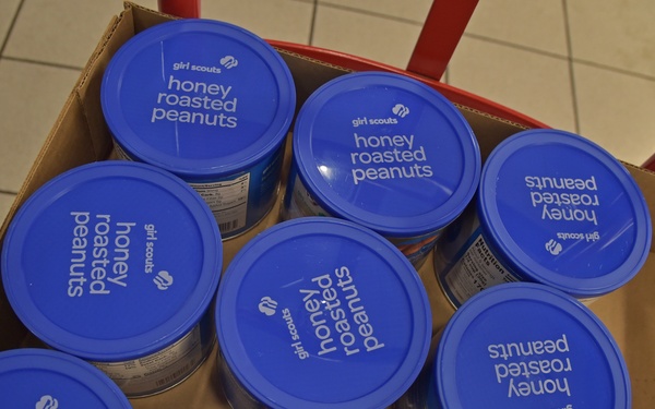 A cardboard box contains multiple canisters of Girl Scouts Honey Roasted Peanuts with blue lids, viewed from above. The canisters are packed tightly together in the box, which appears to be sitting on a red cart or dolly on a tiled floor. An additional canister is partially visible at the bottom right edge of the frame.