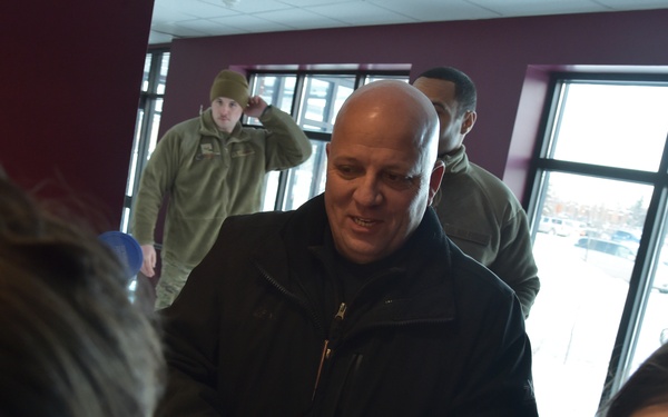 A bald man in a black jacket is interacting with children in what appears to be a building lobby or entrance area. He is holding a blue container, possibly a donation canister, and appears to be engaged in a charitable or community event. Several other adults are visible in the background near glass entrance doors.