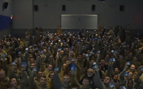 A large group of military personnel, predominantly in camouflage uniforms, are seated in an auditorium or theater-style venue. The majority of attendees are raising small blue cards or booklets in the air, suggesting a coordinated group activity or response. The crowd fills most of the visible seating area, indicating a significant gathering or briefing event.