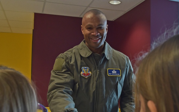 A smiling military aviator in an olive green flight suit is interacting with two young girls, handing them a small container with a blue lid. The setting appears to be an indoor facility with maroon and yellow walls. The interaction appears friendly and positive, suggesting a community outreach or school visit event.