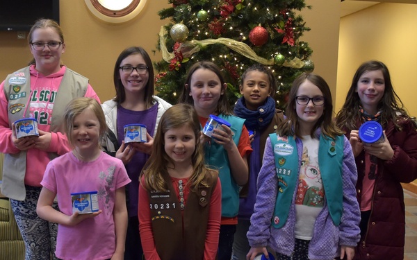 A group of approximately eight girls, several wearing Girl Scout vests with badges and troop number '20231' visible, pose together in what appears to be a lobby or community center decorated for Christmas. Most of the girls are holding small round containers with blue lids, likely donation canisters or product containers associated with a Girl Scout activity. A decorated Christmas tree is visible in the background.