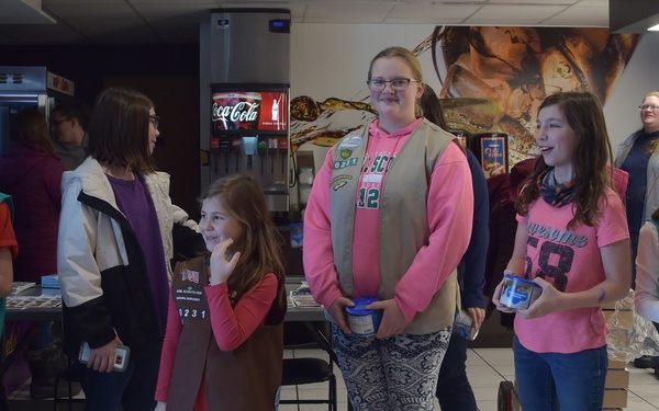 A group of Girl Scouts, identifiable by their green vests with badges, are gathered in what appears to be a movie theater lobby or concession area. Several of the girls are holding small blue containers, possibly donation cans or cookie money boxes. The group includes children of varying ages accompanied by at least one adult chaperone.