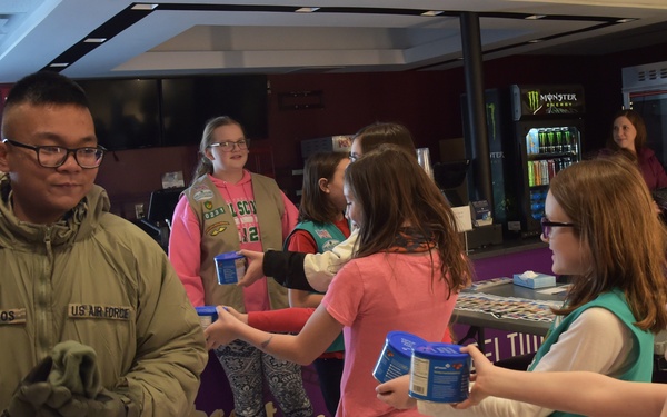 Girl Scouts are handing out items, likely cookie tins or donation containers, to U.S. Air Force personnel in what appears to be a movie theater lobby. A uniformed airman with the name tag 'Oliveros' is visibly smiling while receiving an item from one of the scouts. The scene depicts a community outreach or appreciation event between Girl Scouts and military members.